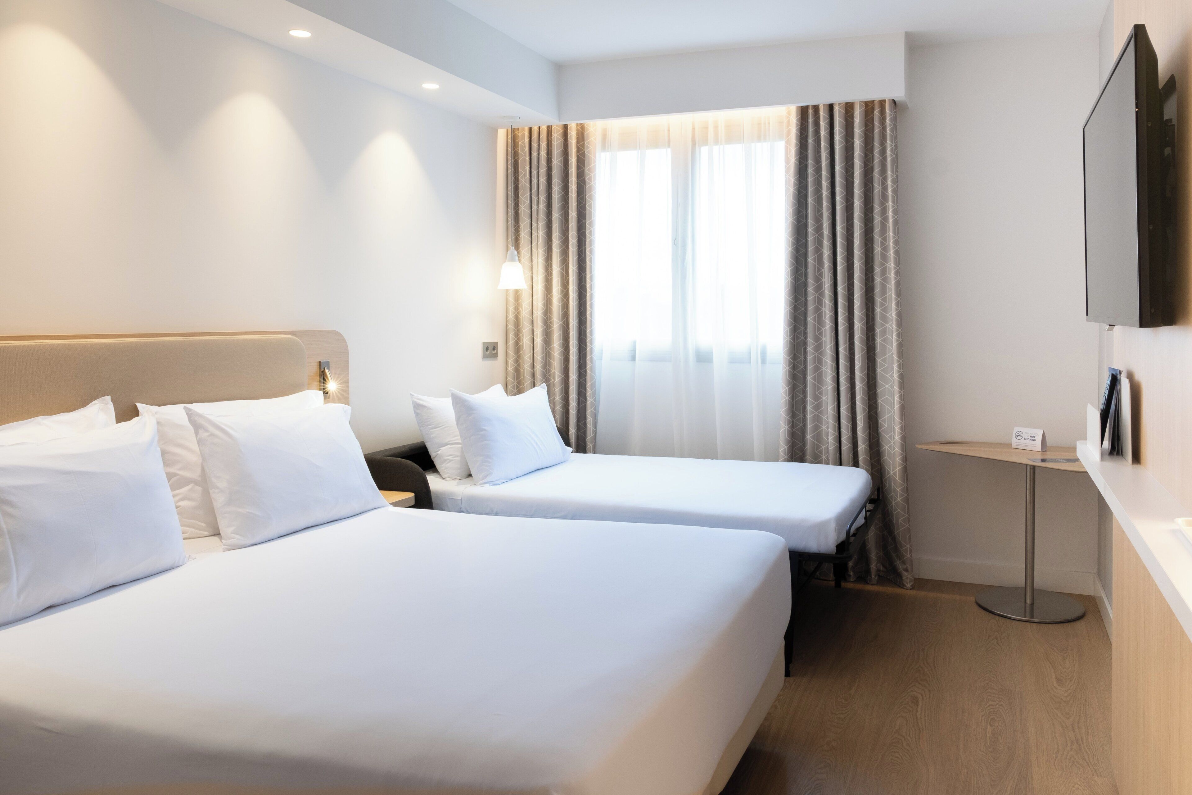 A hotel room with two beds, a TV mounted on the wall, a bedside table, and a window with curtains.