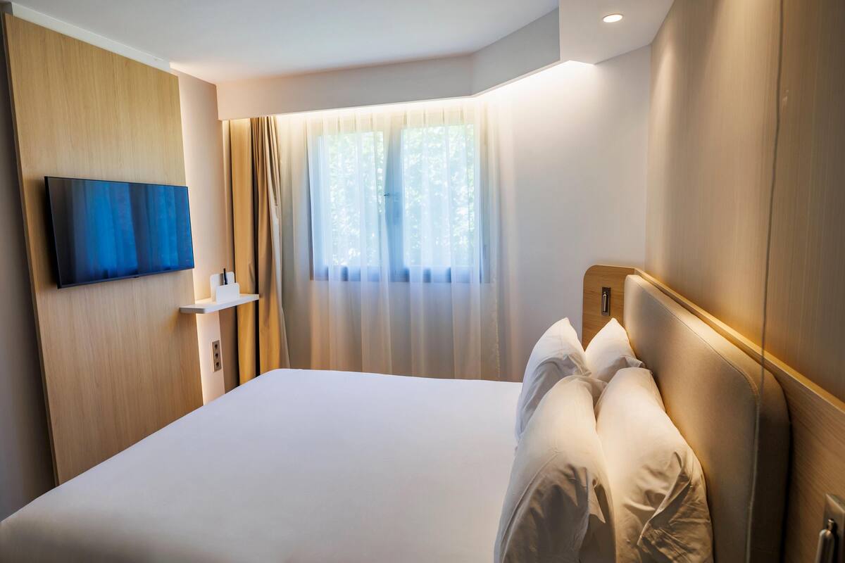 A hotel room with a large bed, a mounted TV, a bedside table, and a window with curtains.