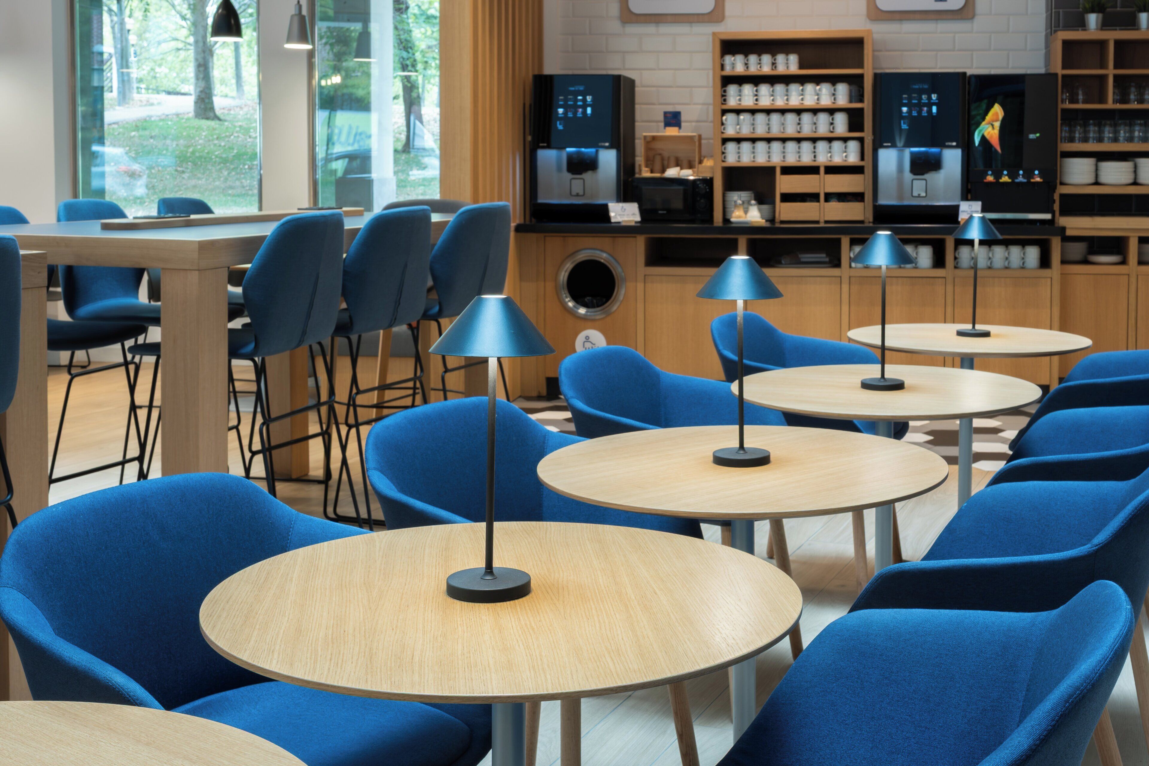 Un café moderno con mesas redondas y sillas azules, una máquina de café y una estantería con tazas y vasos.