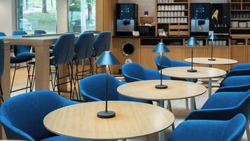 Un café moderno con mesas redondas y sillas azules, una máquina de café y una estantería con tazas y vasos.
