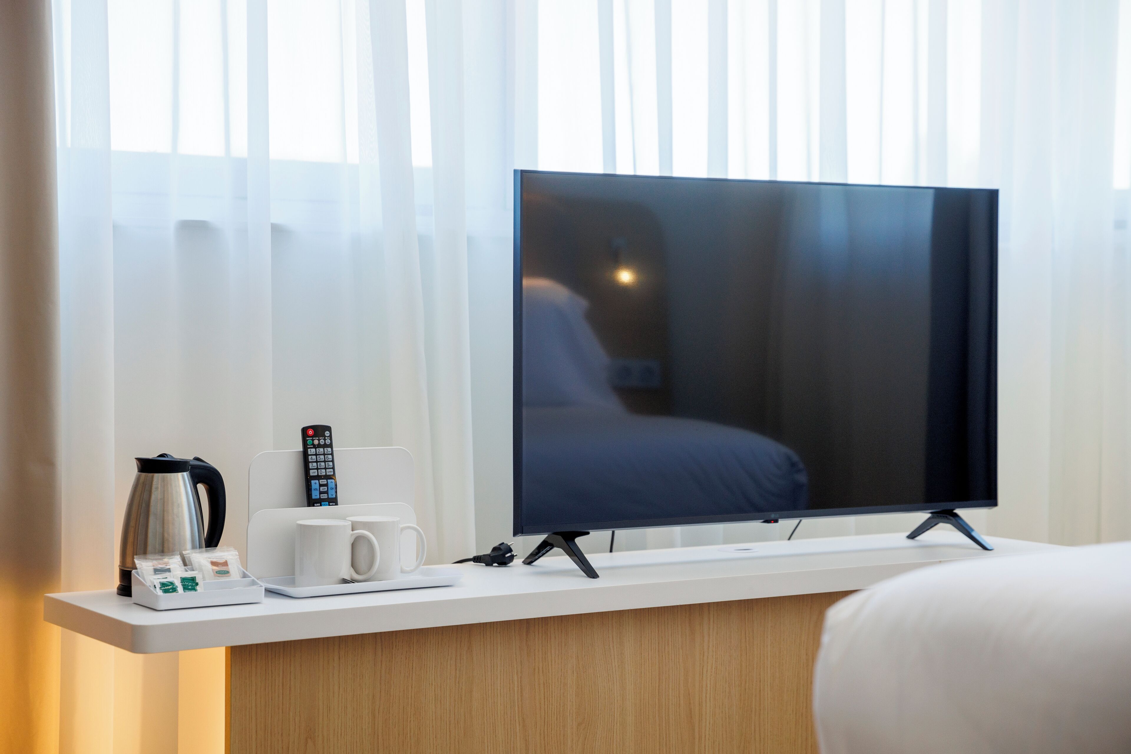 Habitación de hotel con televisor de pantalla plana sobre un pedestal, cafetera, control remoto y bandeja con bolsitas de té y una taza.