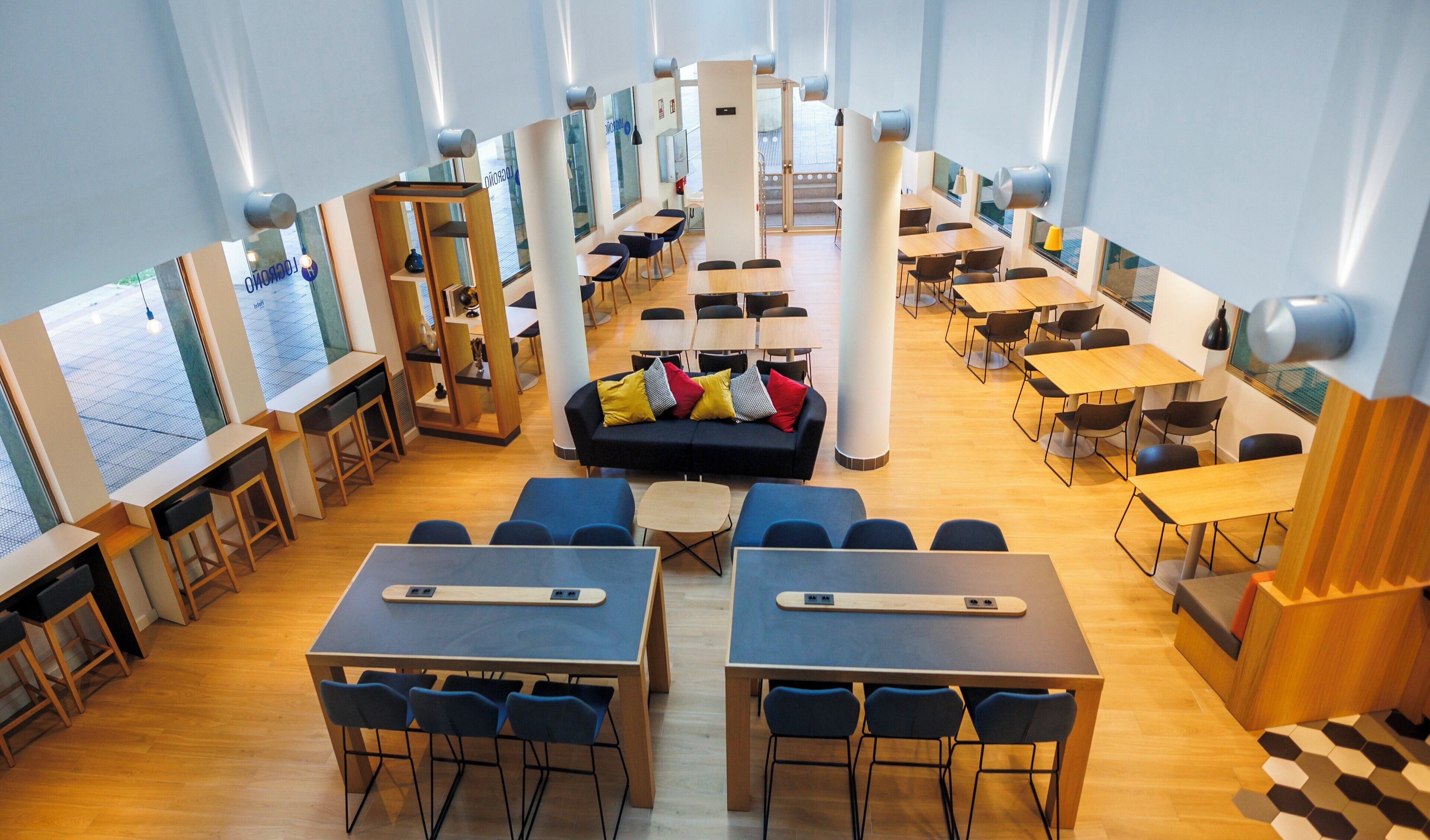 A modern library with multiple tables and chairs, a central sofa, and large windows providing natural light.