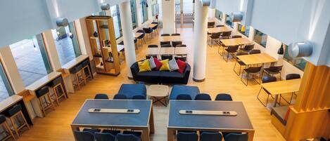 A modern library with multiple tables and chairs, a central sofa, and large windows providing natural light.