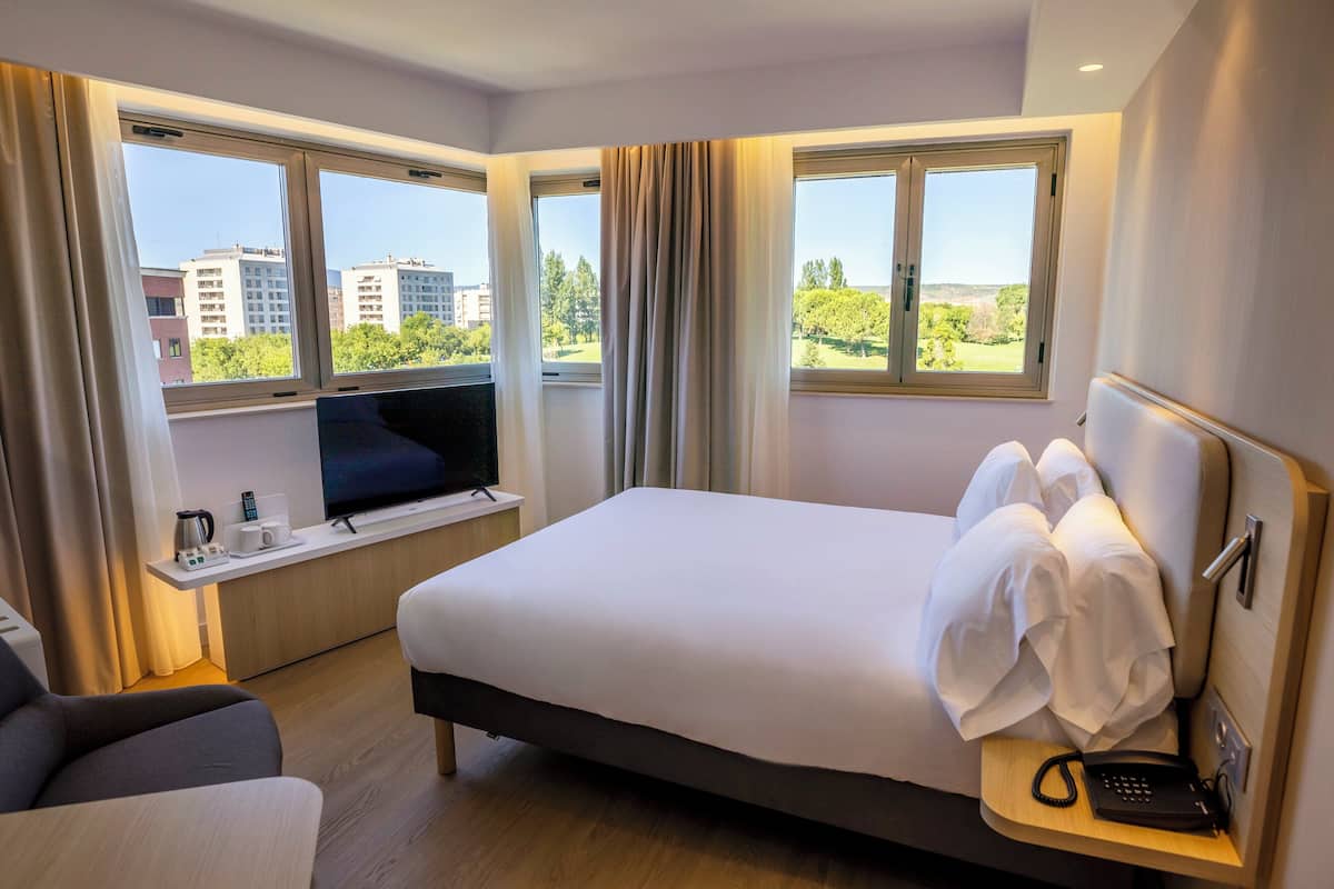 Habitación de hotel con una cama grande, un televisor sobre un mueble bajo, un escritorio con teléfono y vistas a edificios y árboles a través de las ventanas.
