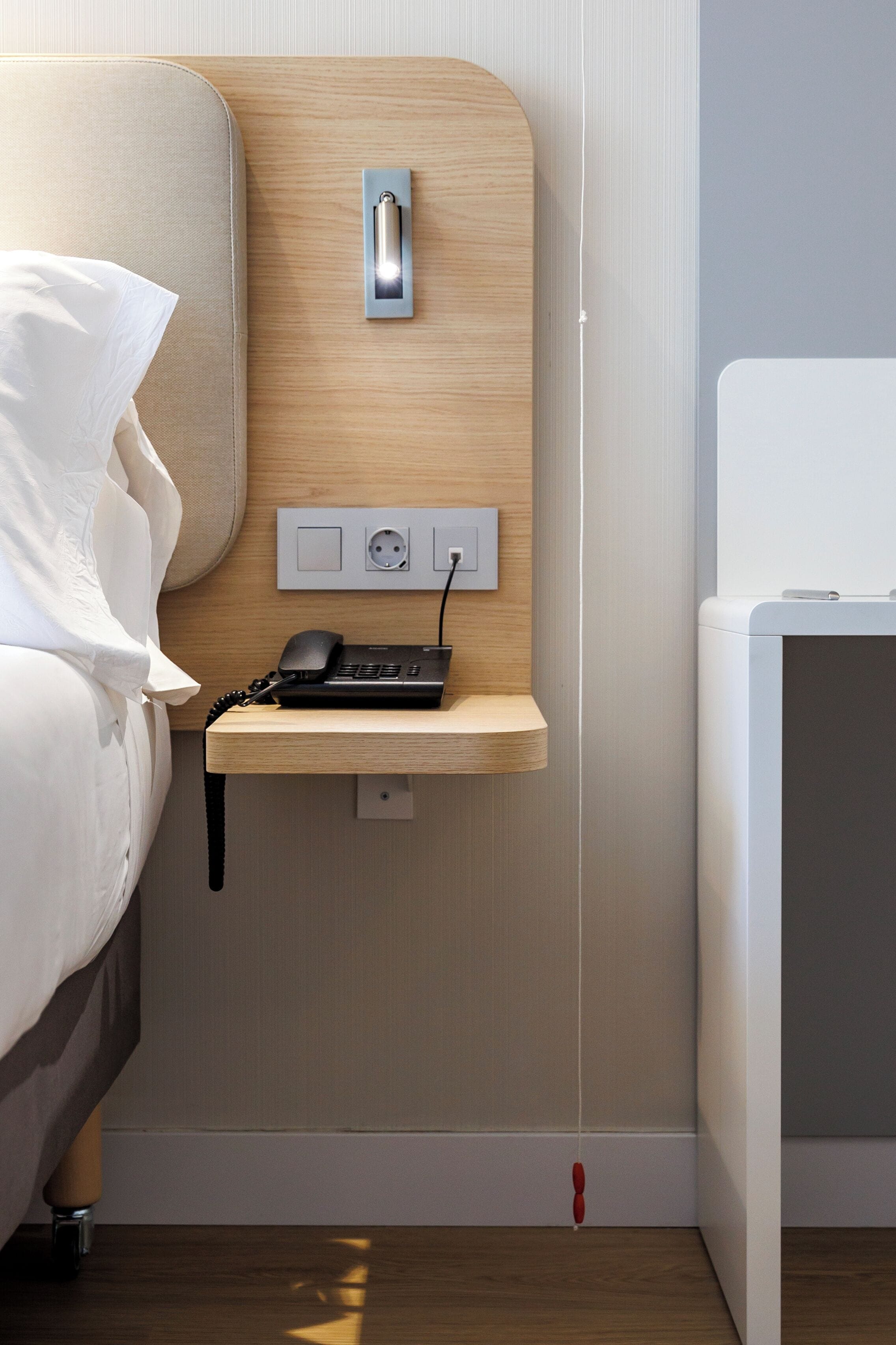 A hotel room with a bed, a bedside telephone, a power outlet, and a small wooden shelf.