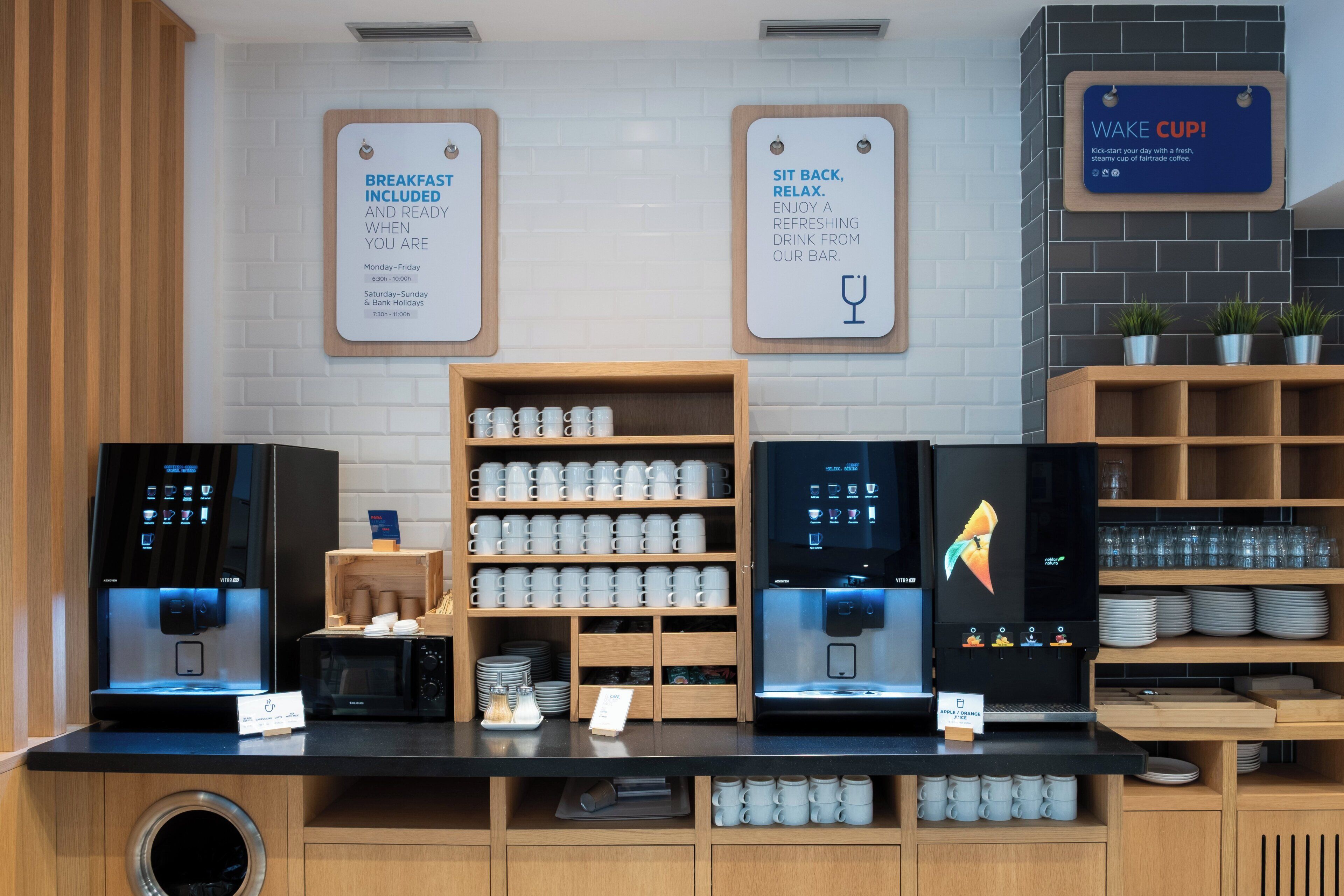 A coffee station with multiple coffee machines, a sign indicating breakfast is included, and a sign encouraging guests to sit back and relax.