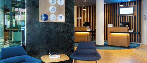 Un vestíbulo de hotel moderno con mostrador de recepción, dos sillas azules, una mesa pequeña con un libro y un letrero en la pared con iconos direccionales.