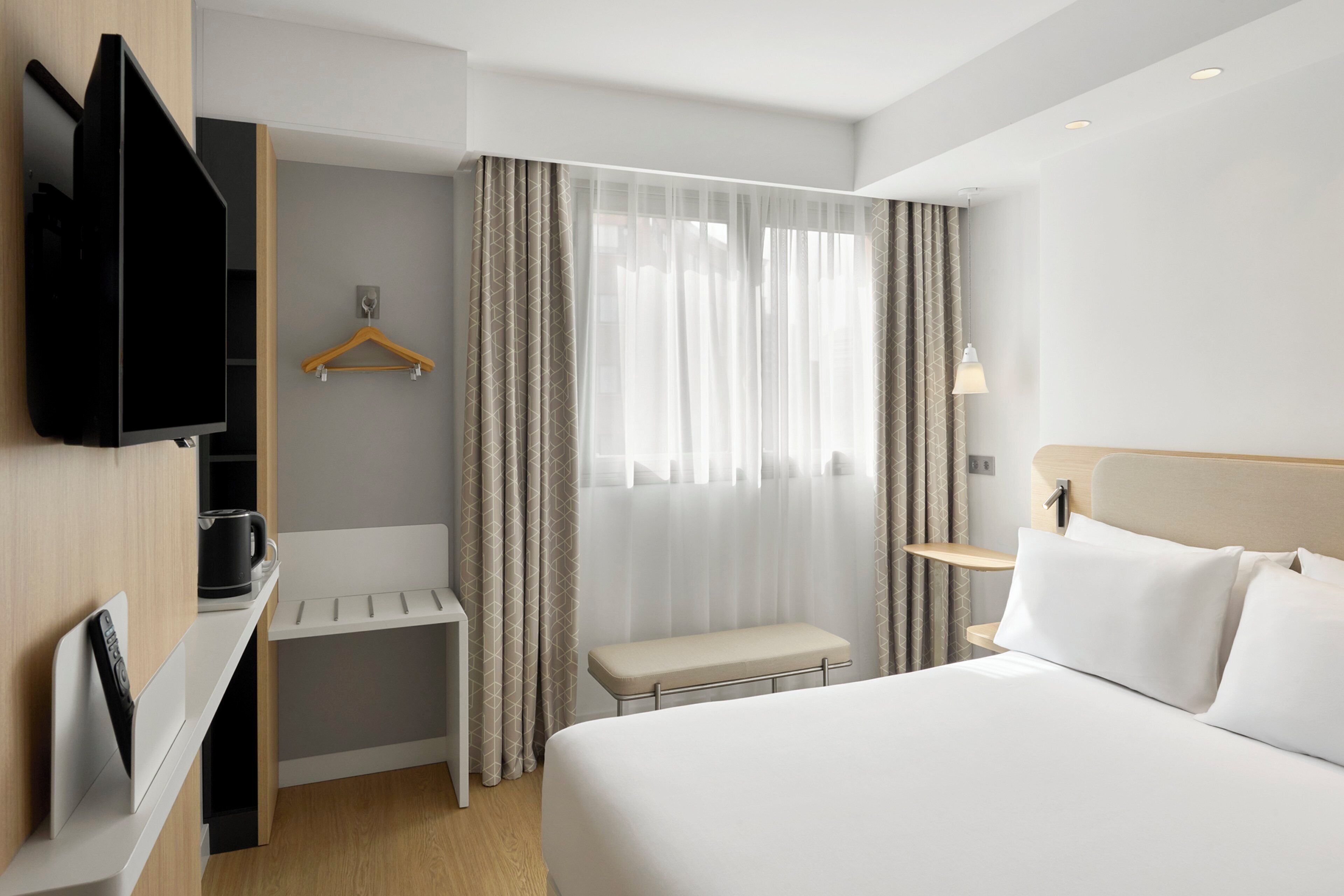 A hotel room with a bed, a TV mounted on the wall, a bench, a bedside table, and a window with curtains.
