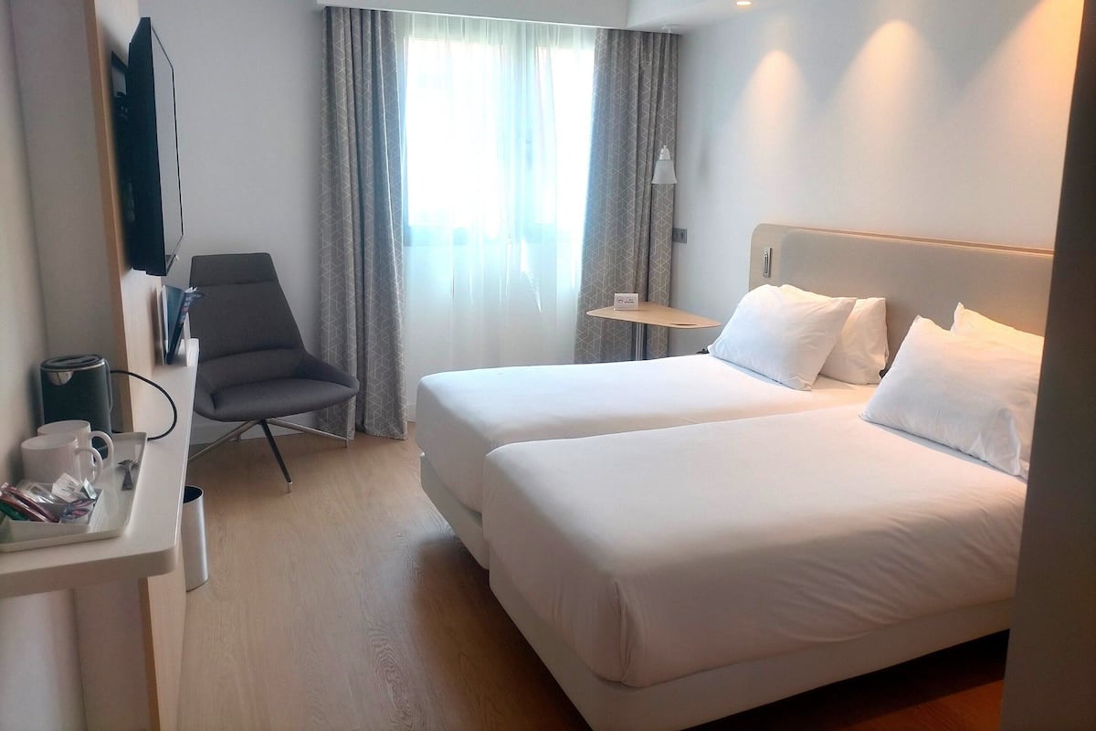 Una habitación de hotel con dos camas, un escritorio con silla, televisión y ventana con cortinas.