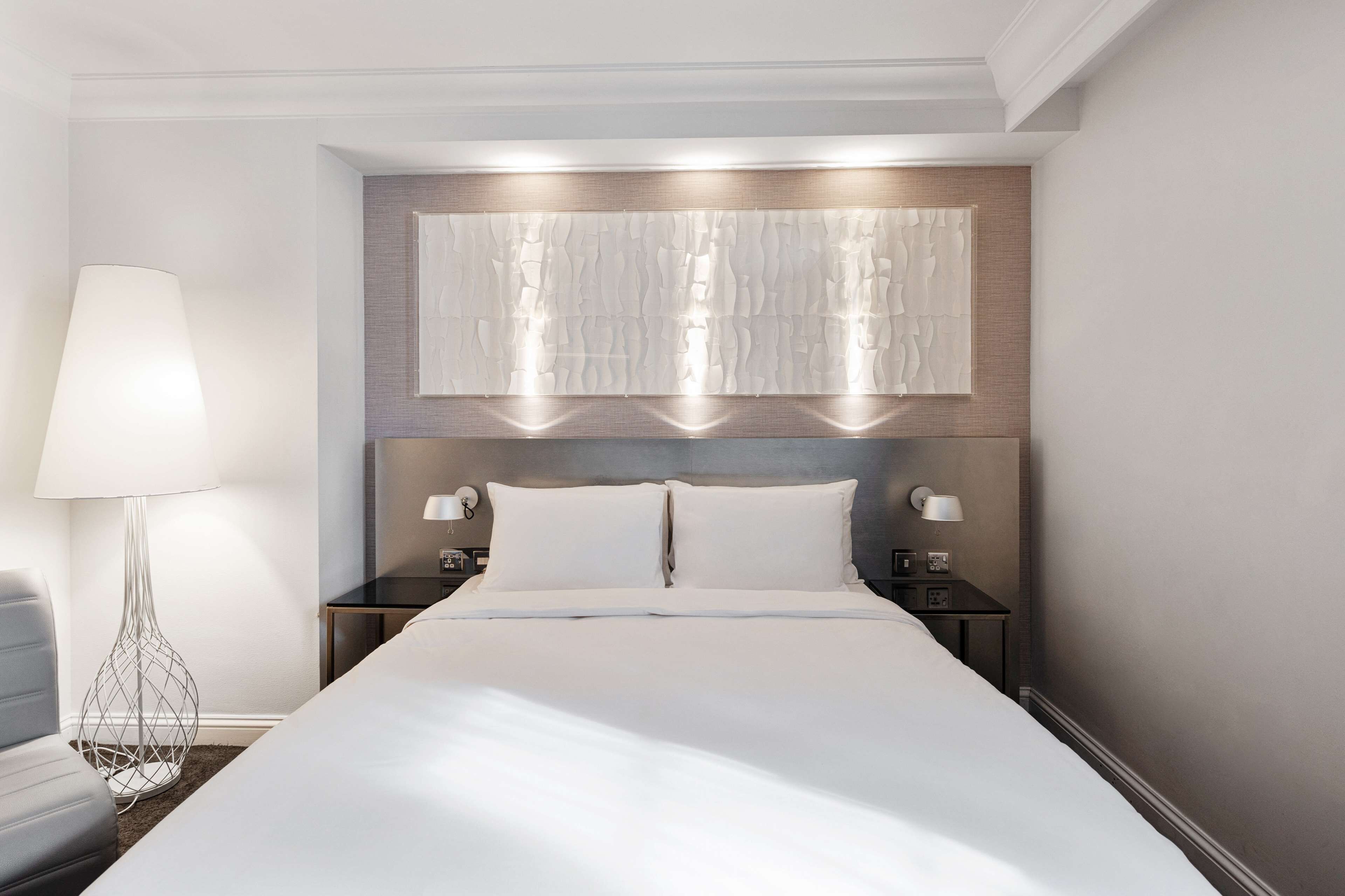 Une chambre d’hôtel moderne avec un grand lit, deux tables de chevet, un luminaire fixé au mur et un panneau décoratif au-dessus du lit.