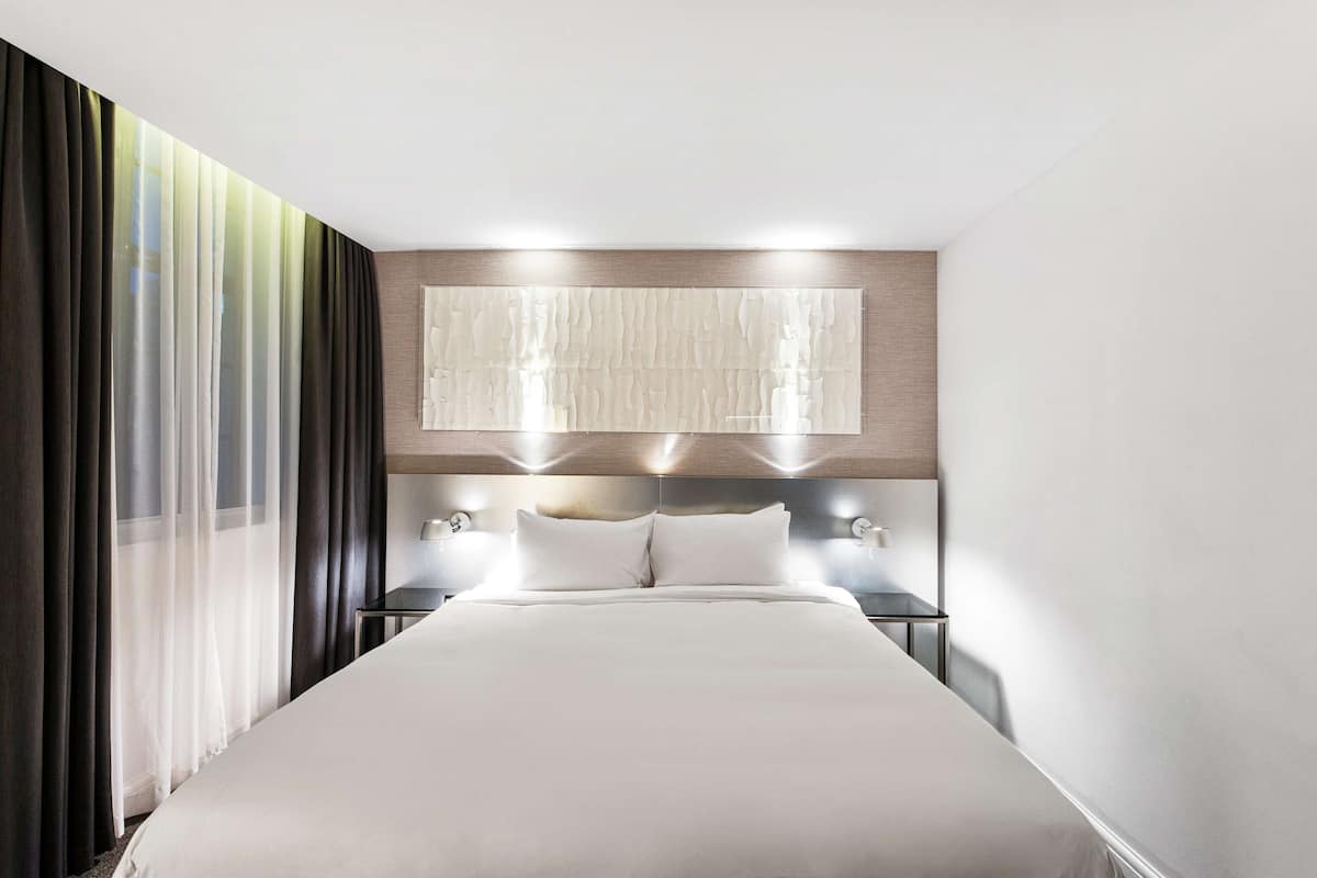 A modern hotel room with a large bed, a headboard with integrated lighting, a nightstand with a lamp, and sheer curtains on the window.