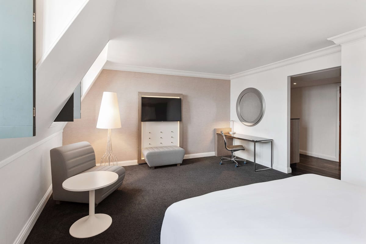 A hotel room with a bed, a desk, a chair, a sofa, a lamp, and a TV.