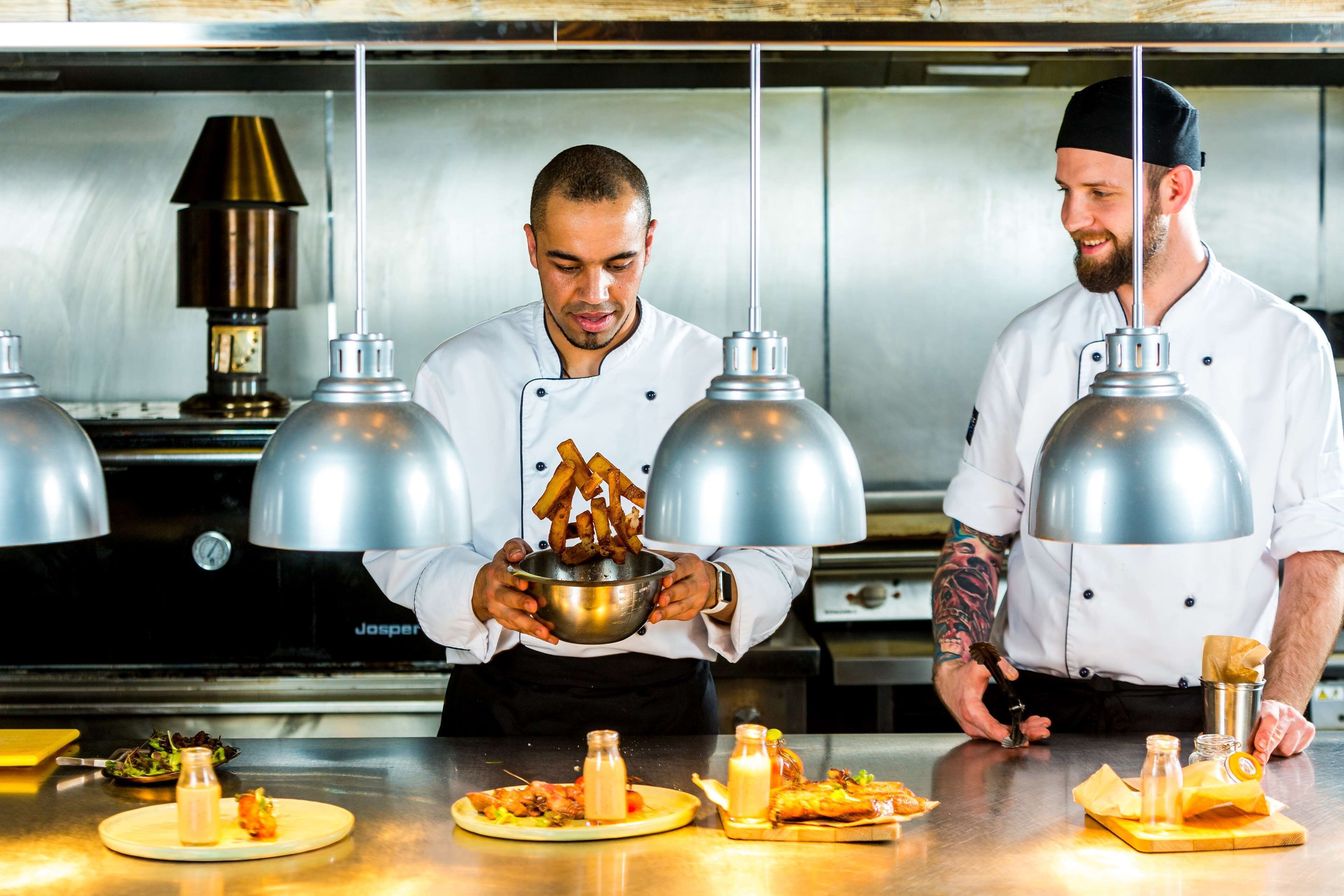 Des chefs travaillent dans une cuisine professionnelle équipée d’appareils en acier inoxydable, avec une grande marmite et divers ingrédients disposés sur le plan de travail.