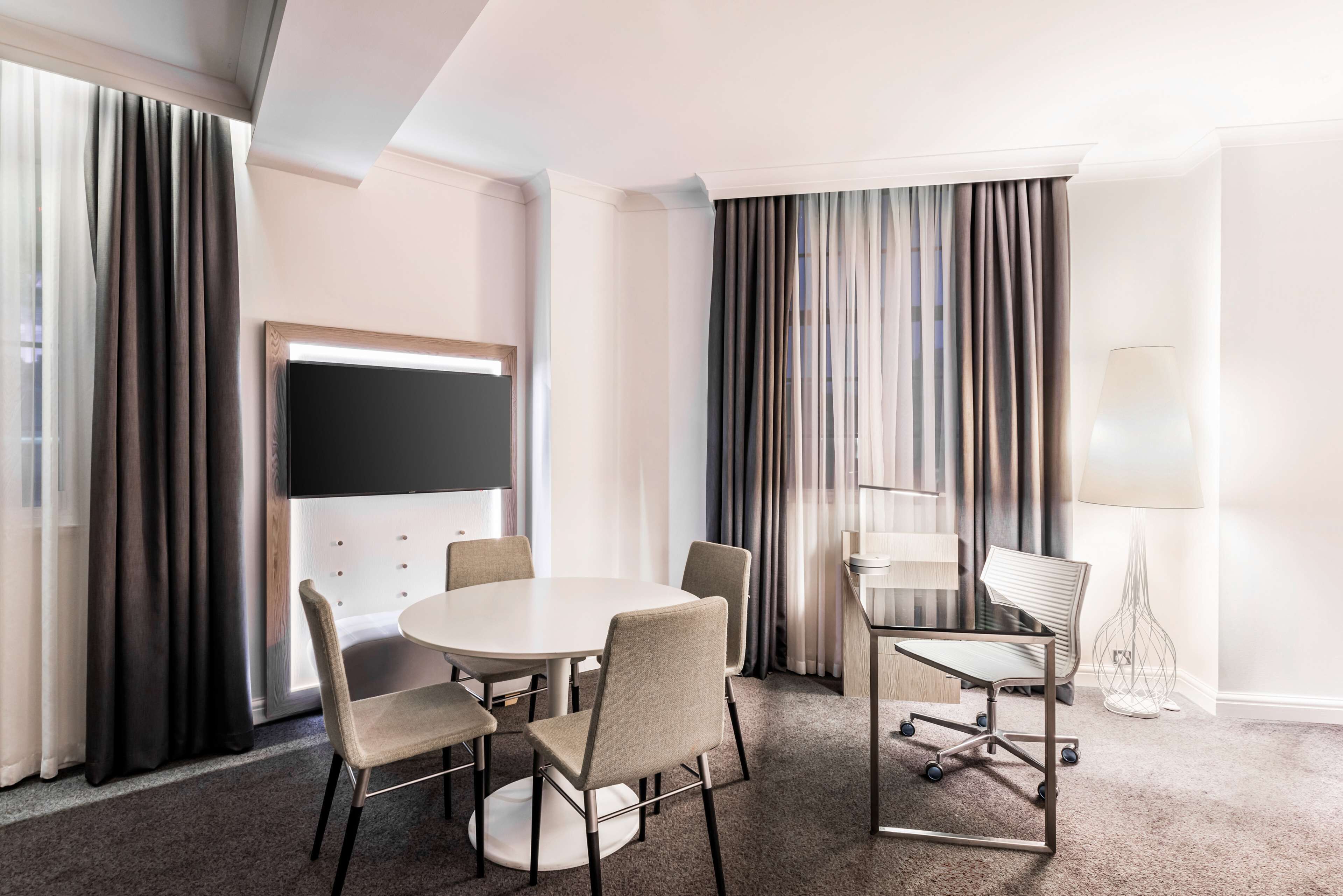 Ein Hotelzimmer mit rundem Tisch, vier Stühlen, einem Stehpult, einem Fernseher und einer Lampe.