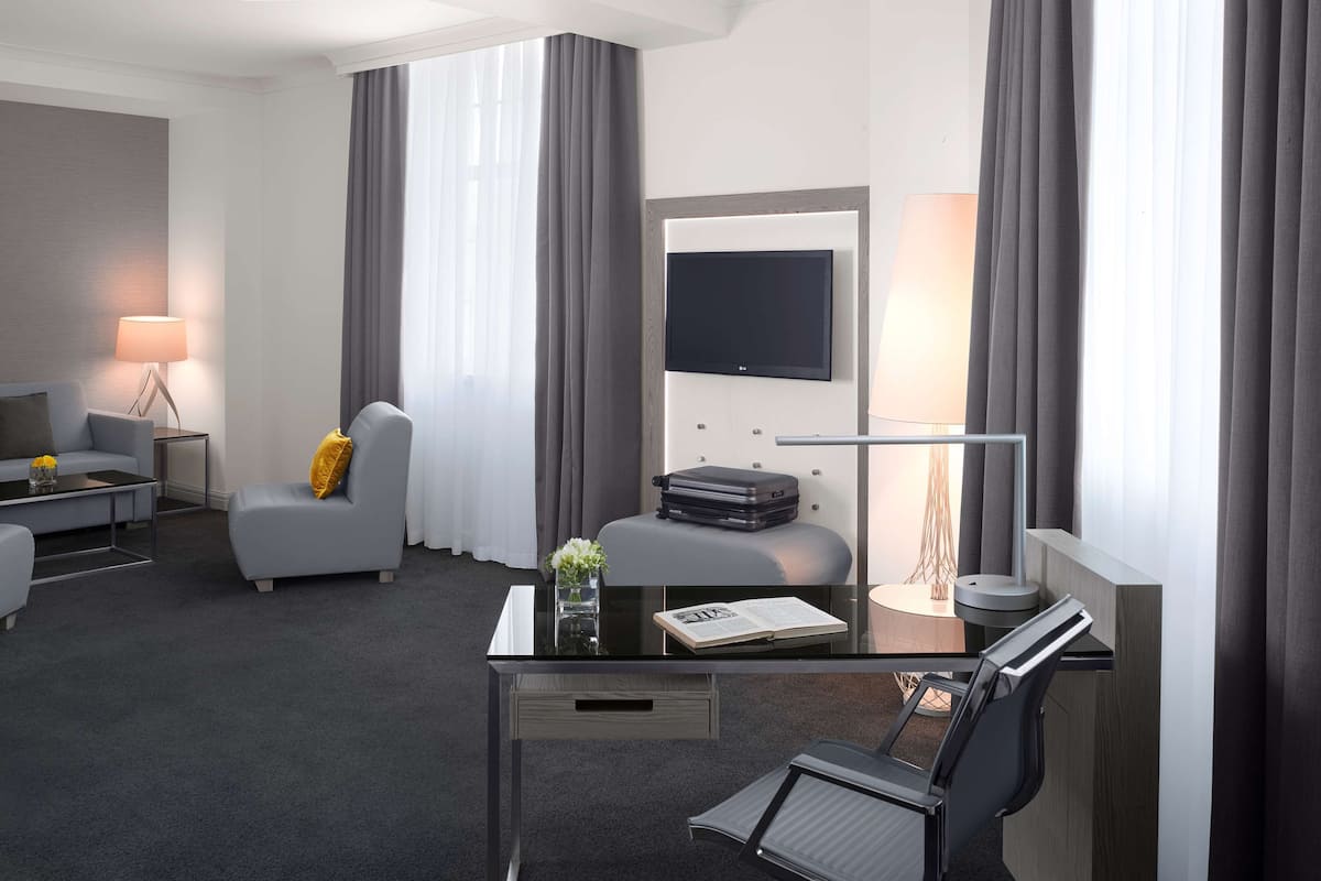 A modern hotel room with a grey sofa, a small armchair, a desk with a chair, a flat-screen TV, and a lamp.
