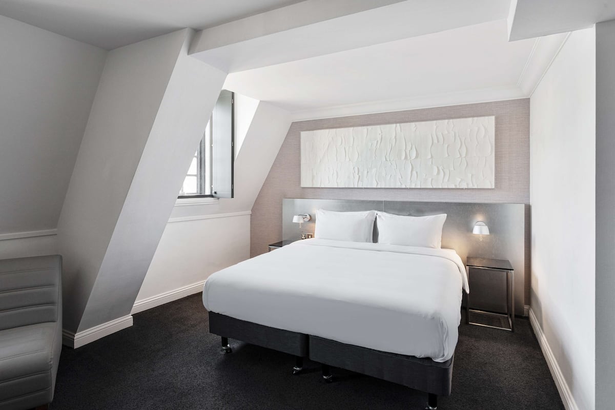 A modern hotel room with a large bed, a headboard with a textured design, two bedside tables with lamps, and a window providing natural light.