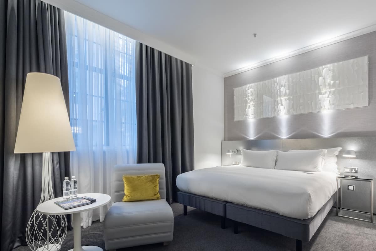 A modern hotel room with a large bed, a small armchair, a bedside table with a lamp, and a view of the city through large windows.