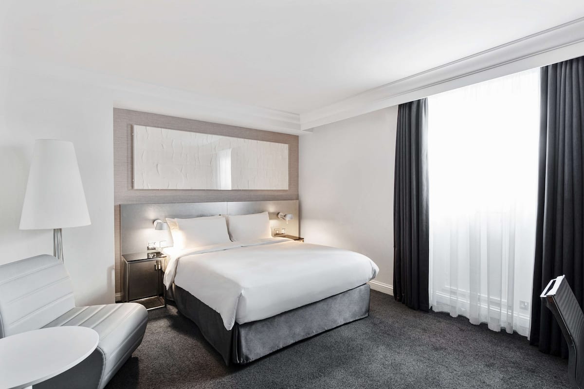 A hotel room with a bed, bedside table, lamp, and a large window with curtains.