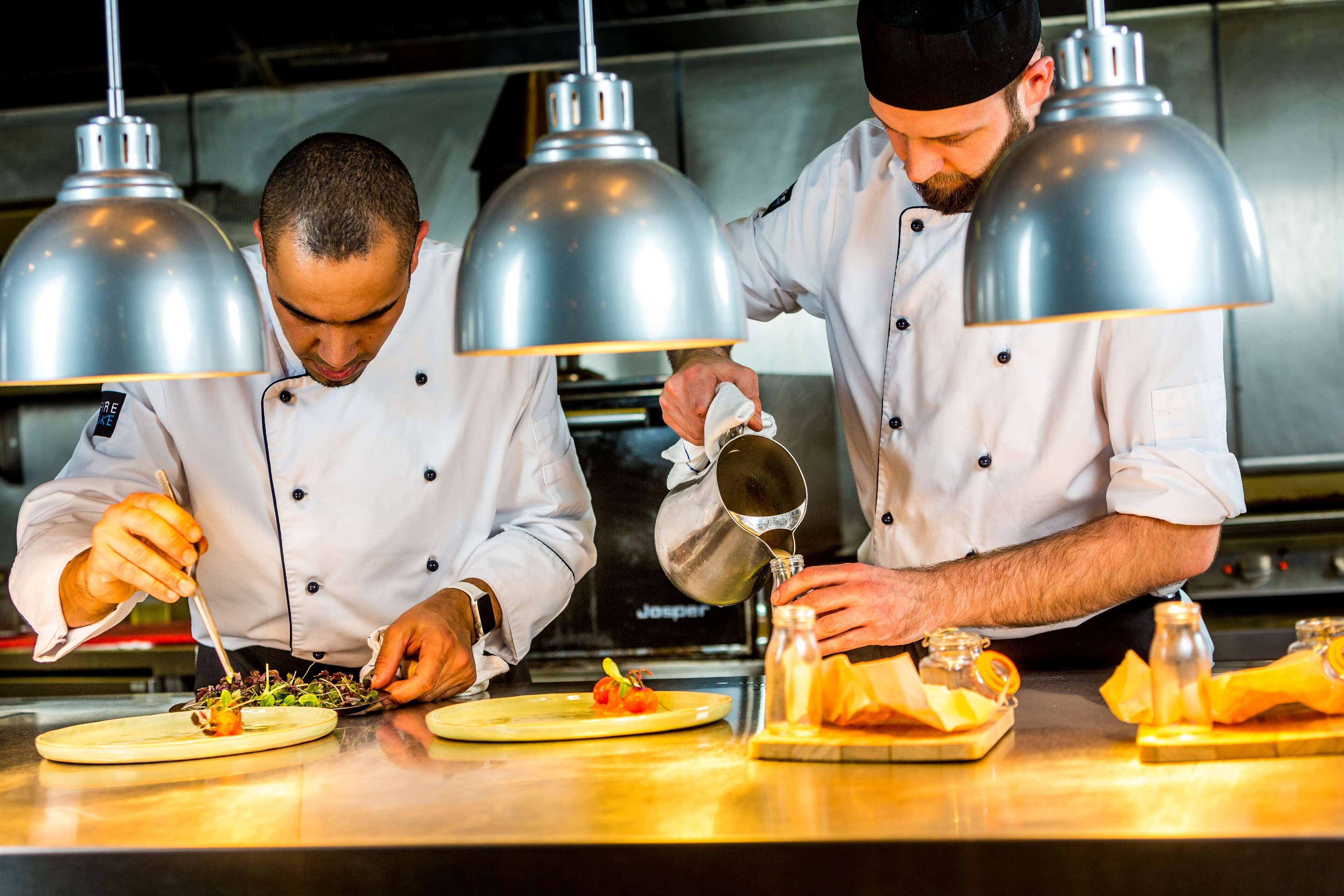 Chefs preparing food in a professional kitchen with stainless steel surfaces and pendant lighting.
