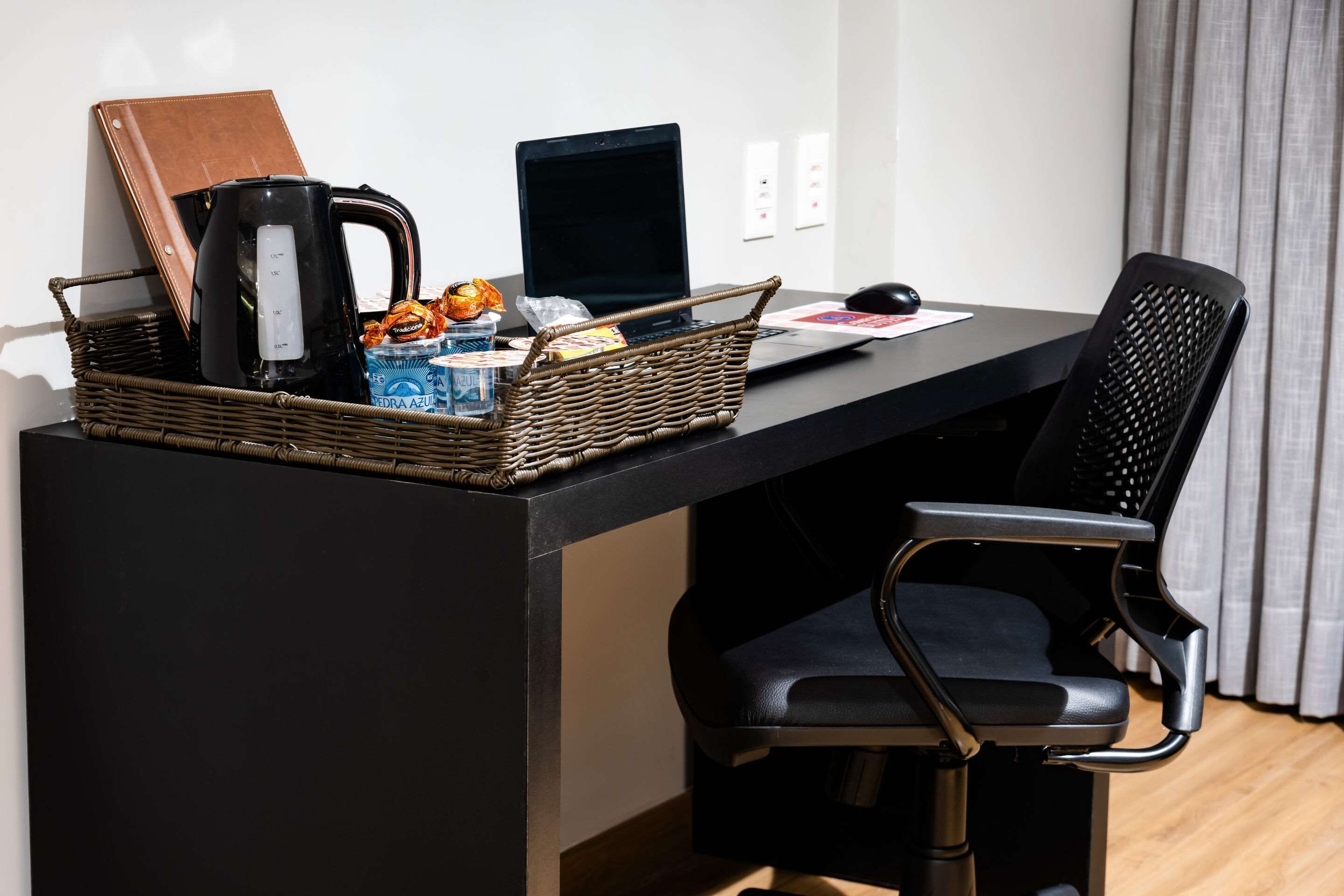 Een moderne werkplek met een laptop, een koffiezetapparaat, een rieten mand met snacks, en een lederen notitieboek op een zwart bureau.