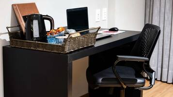 Un espacio de trabajo moderno con una computadora portátil, una cafetera, una cesta de mimbre con aperitivos y una libreta de cuero sobre un escritorio negro.