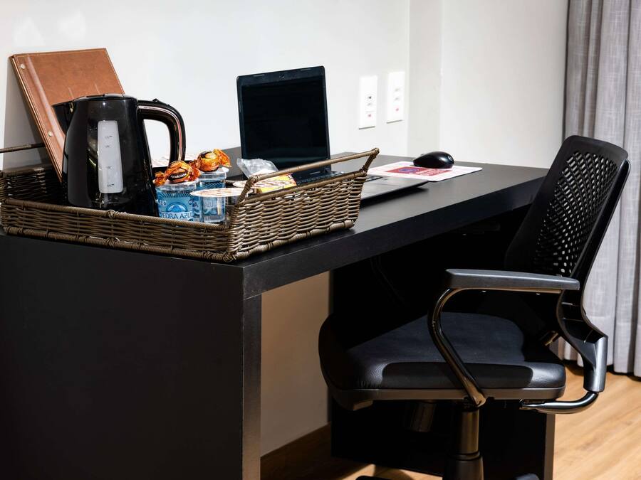 Un espacio de trabajo moderno con una computadora portátil, una cafetera, una cesta de mimbre con aperitivos y una libreta de cuero sobre un escritorio negro.