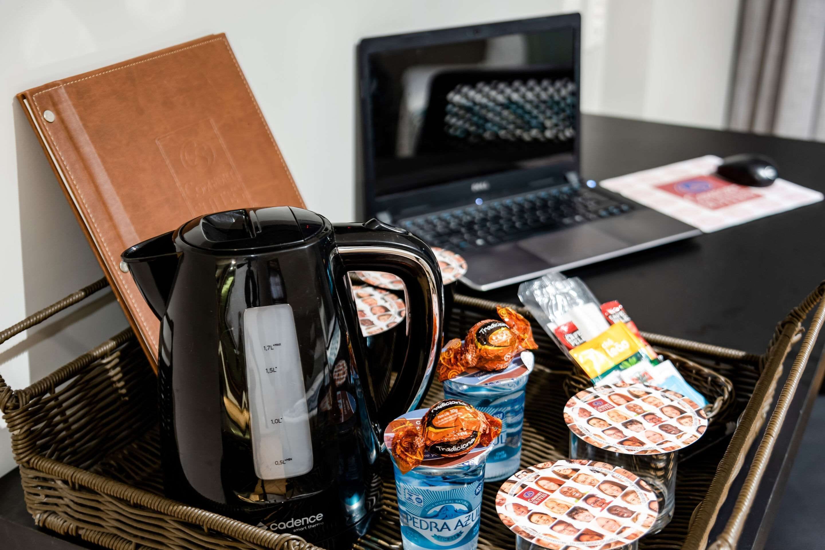 A laptop, a coffee maker, a laptop mouse, a notebook, and a wicker basket with drinks and snacks.