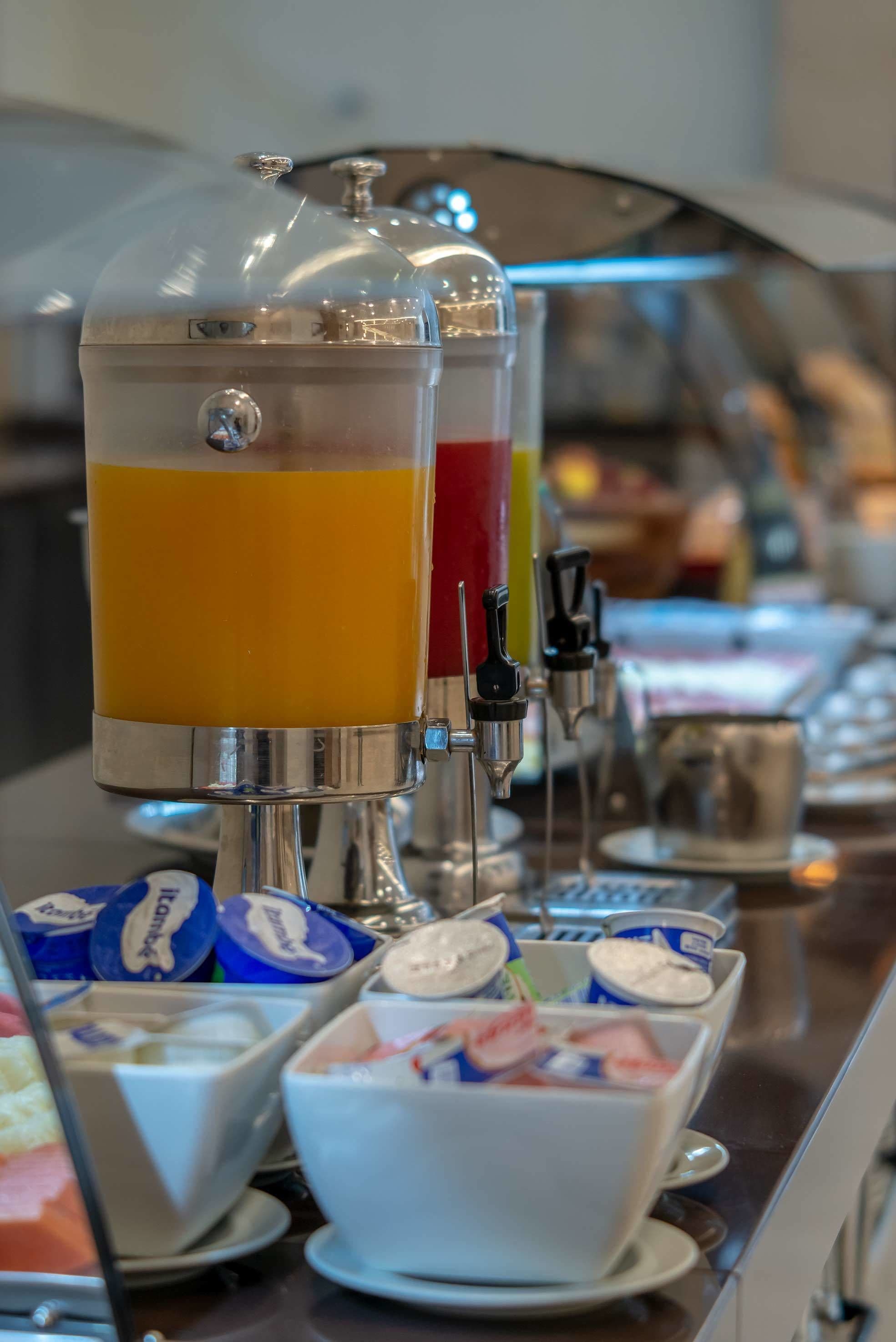 A beverage dispenser with multiple options, including orange juice, and a selection of yogurt cups and bowls.