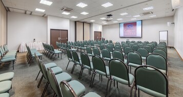 Un salón de conferencias con hileras de sillas verdes, una pantalla de proyección que muestra 'Comfort Suites' y piso alfombrado.