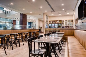 Un restaurante moderno con mesas altas y cabinas, una zona de bar con mostrador y estanterías llenas de botellas, y una pantalla grande en la pared.