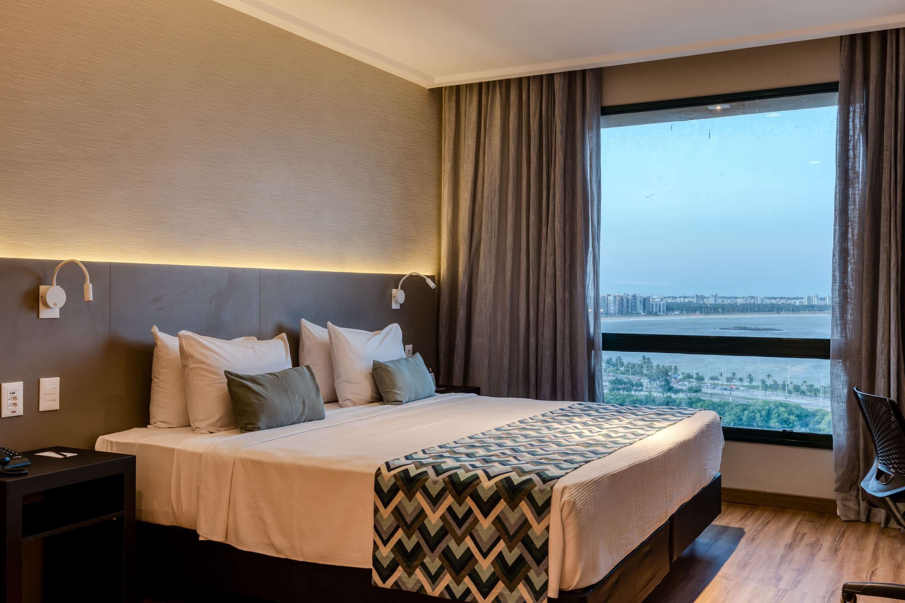 Ein modernes Hotelzimmer mit einem großen Bett, einem Nachttisch, einem Sessel und Blick auf eine Stadtlandschaft durch ein großes Fenster.