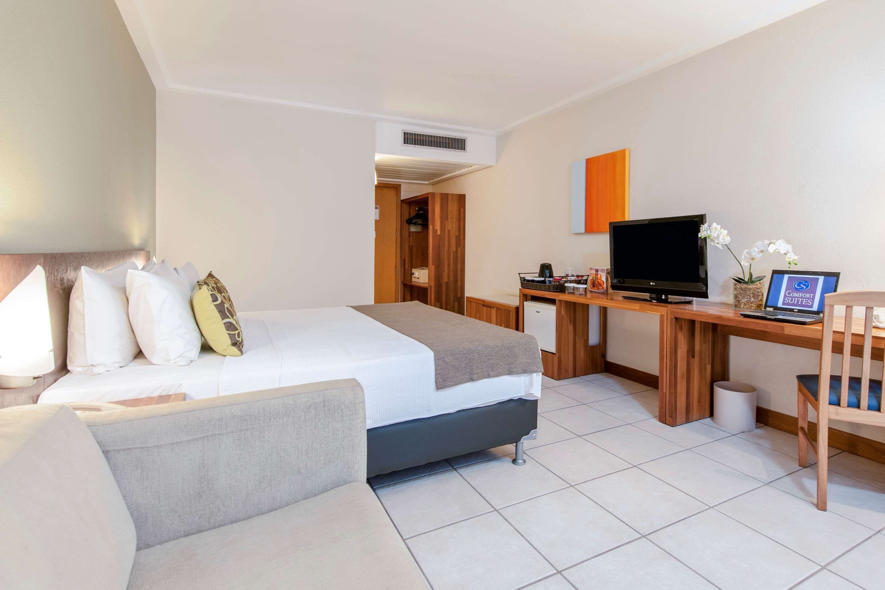 Una habitación de hotel moderna con una cama, un escritorio con computadora, televisor y un pequeño rincón de descanso.