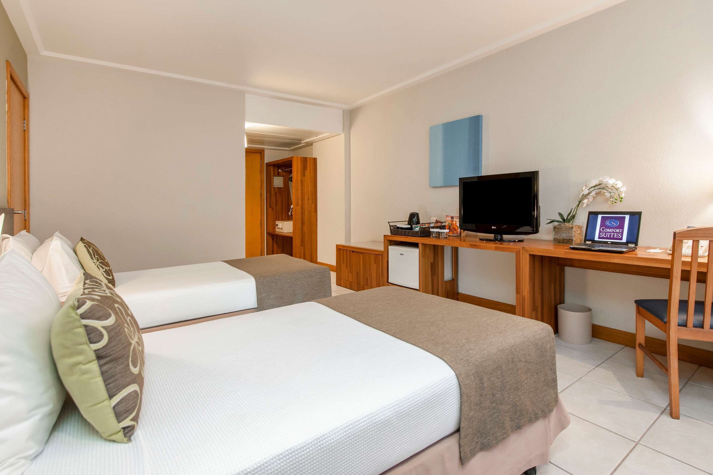 Quarto de hotel com duas camas, uma mesa de escritório, computador, televisão e um pequeno frigorífico.