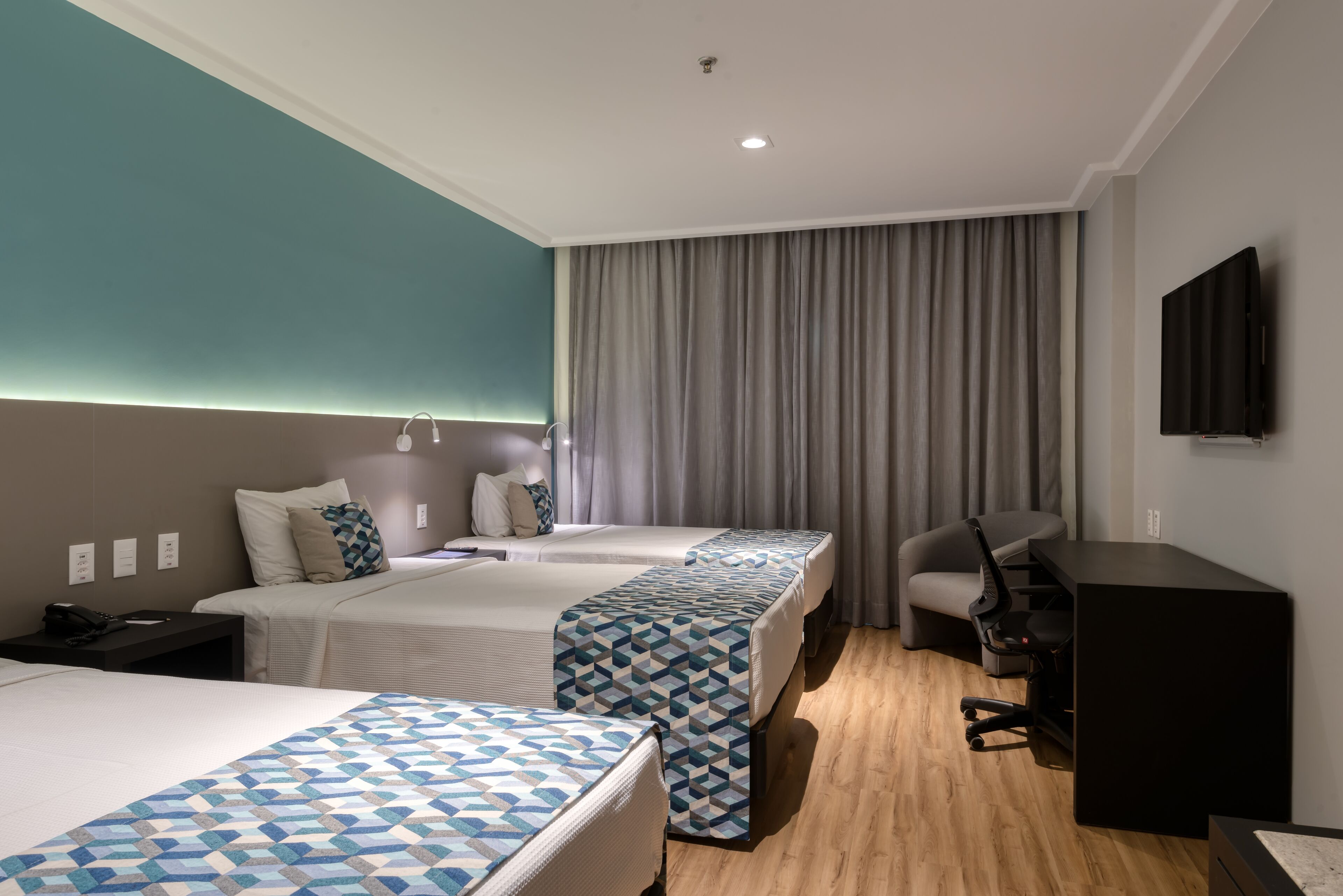 A hotel room with two beds, a desk, a chair, a TV, and a large window with curtains.