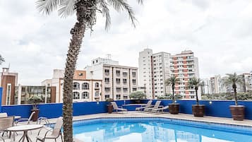 Una piscina en la azotea con sillones de descanso, una palmera y rascacielos al fondo.