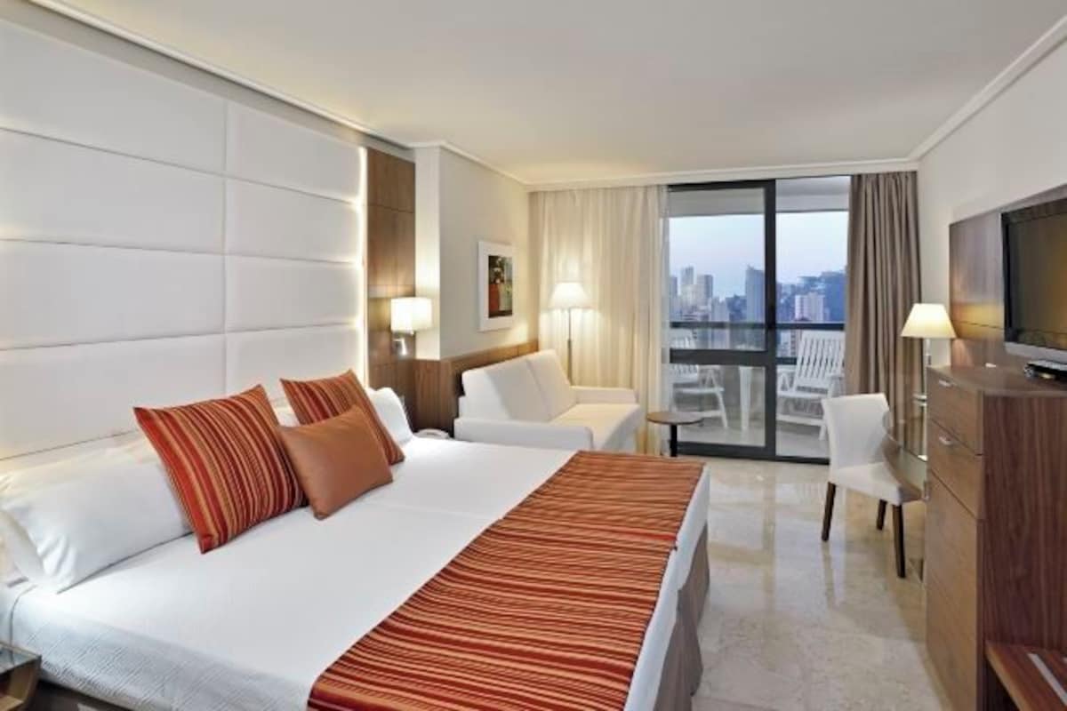 A hotel room with a large bed, a TV, a desk, and a balcony with a city view.