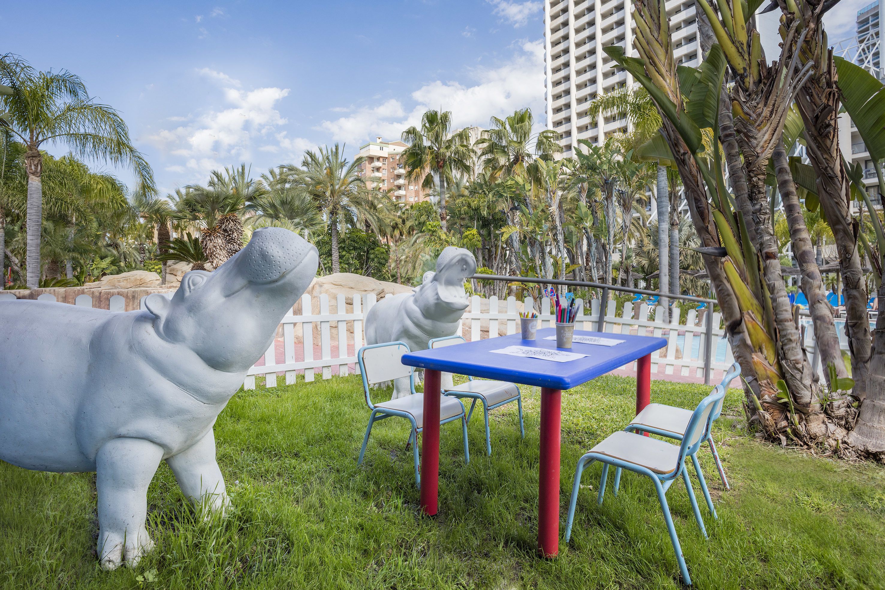 A grassy outdoor area with a white statue of a hippo, a blue table with chairs, and a white picket fence.