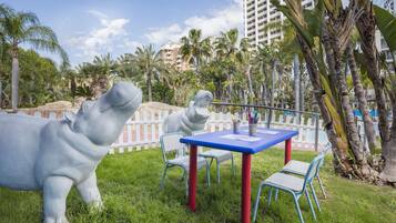 A grassy outdoor area with a white statue of a hippo, a blue table with chairs, and a white picket fence.
