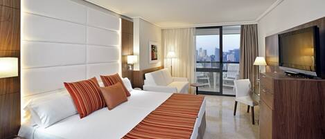 A hotel room with a large bed, a TV, a desk with a laptop, a chair, and a balcony with city views.