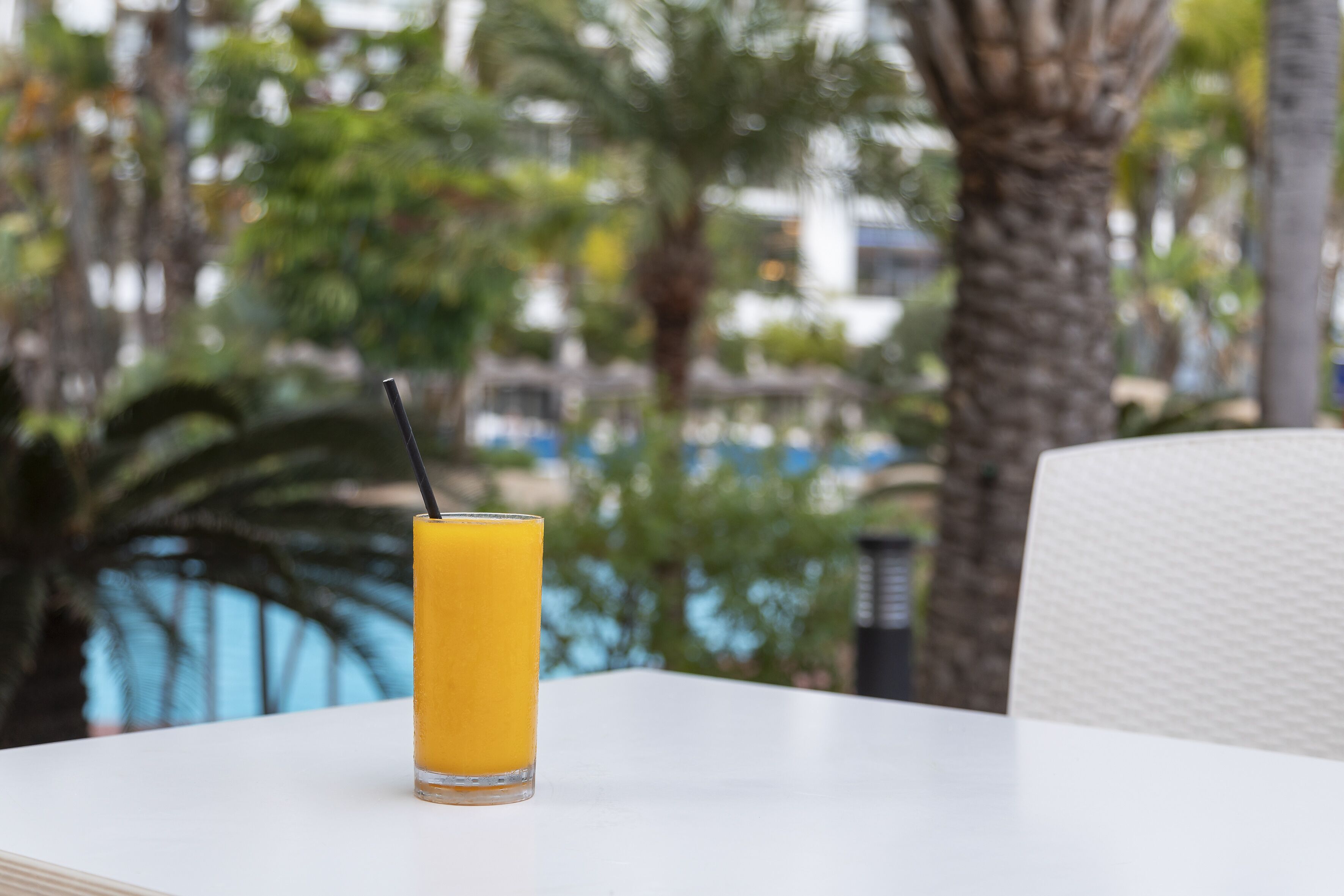 Un vaso de jugo de naranja sobre una mesa blanca, con un fondo desenfocado de palmeras y una piscina.