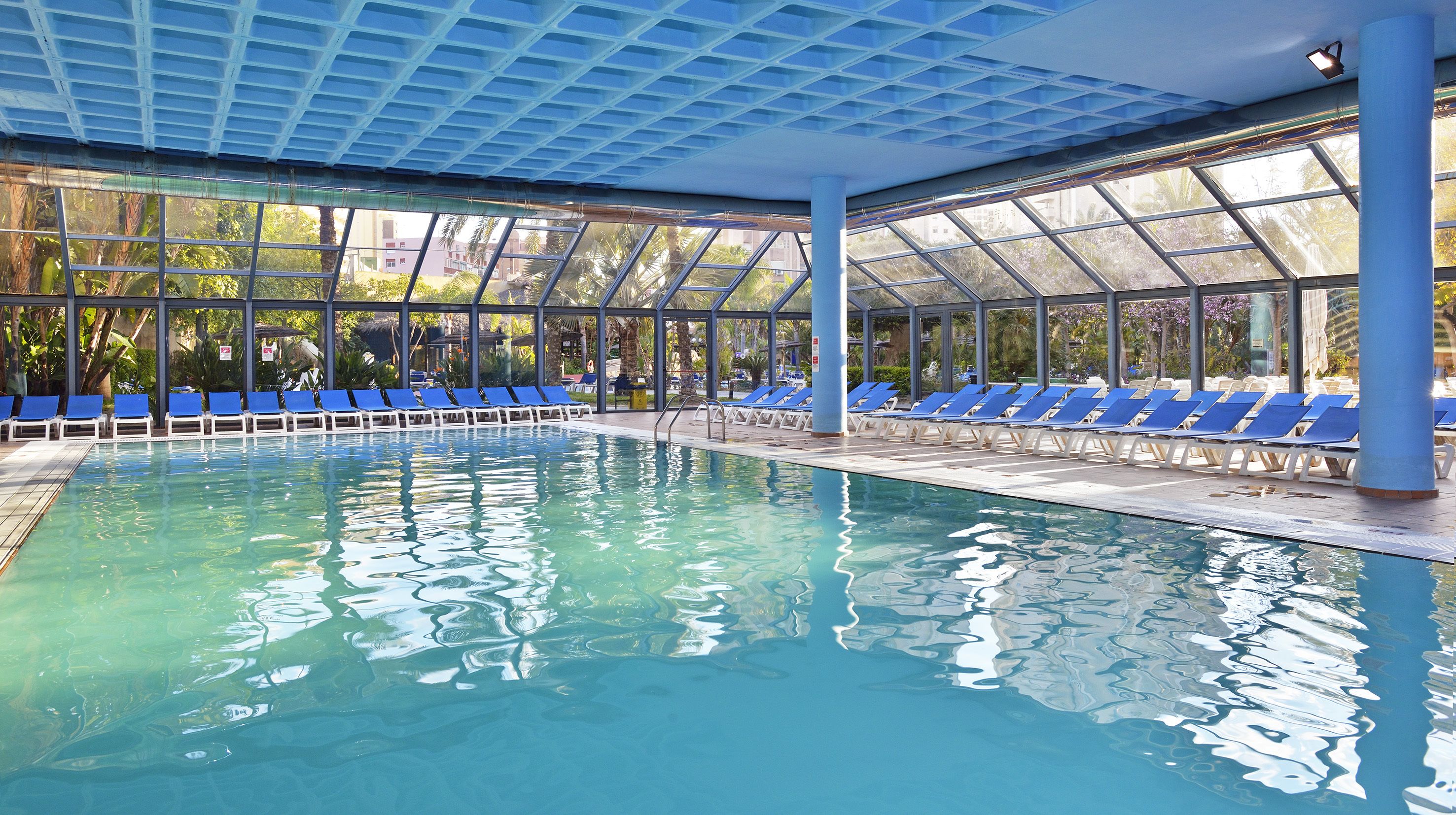An indoor swimming pool with clear blue water, surrounded by blue lounge chairs and a glass ceiling.
