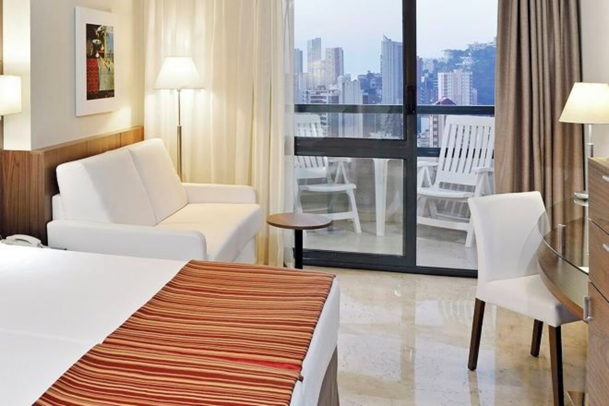A hotel room with a bed, two white armchairs, a small table, and a view of the city through a large window.