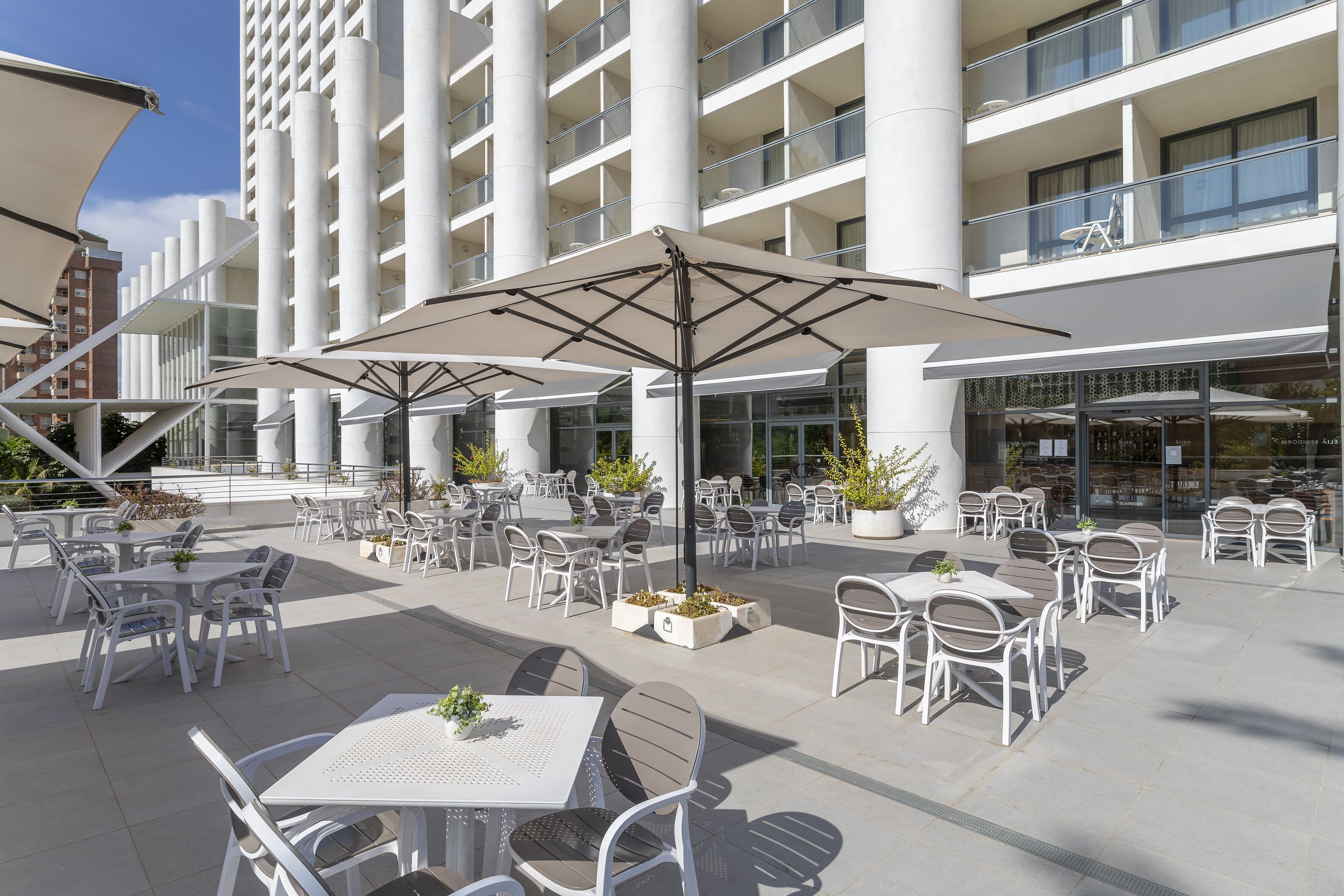 An outdoor patio area with white tables and chairs, umbrellas, and potted plants.