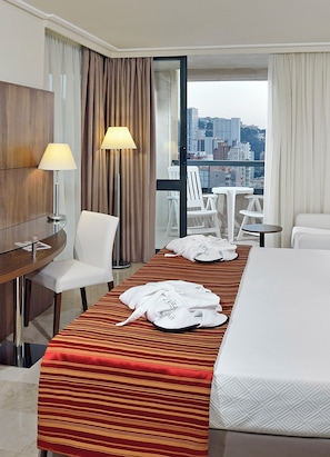 A hotel room with a bed, a desk with a lamp, a chair, a table with chairs, and a view of the city through large windows.