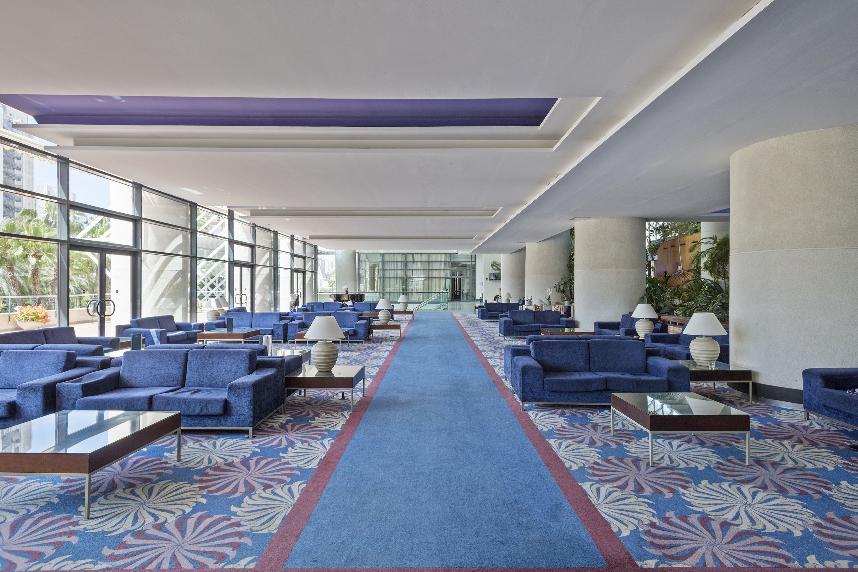 Un vestíbulo de hotel con un pasillo central alfombrado, sofás azules y mesas con lámparas.