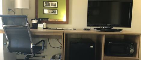 A hotel room with a desk, computer, chair, lamp, and a TV.