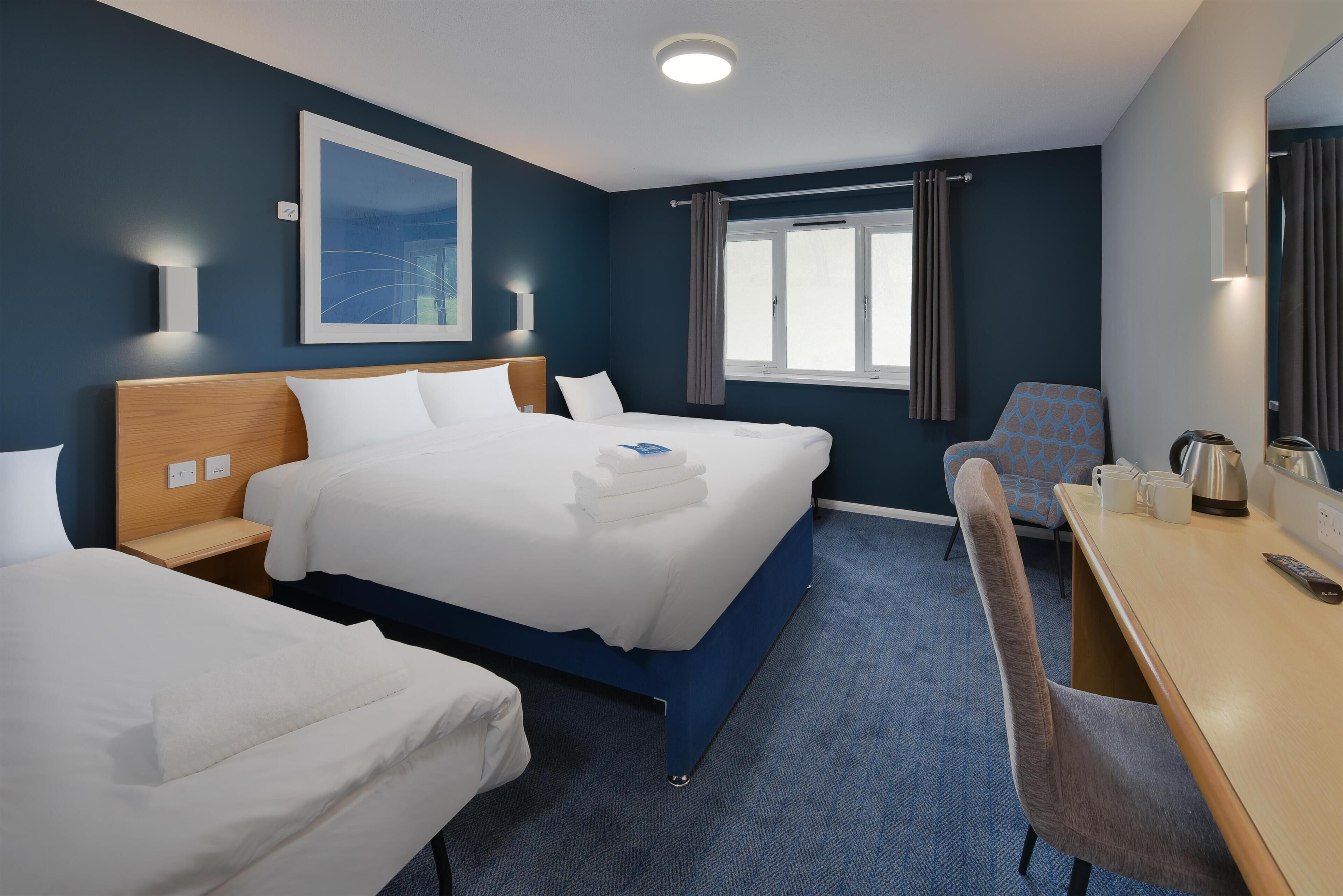A hotel room with two beds, a desk, a chair, a window with curtains, and a painting on the wall.