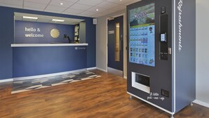 A reception area with a blue wall, a clock, a 'hello & welcome' sign, a Travelodge logo, and a refreshment vending machine.
