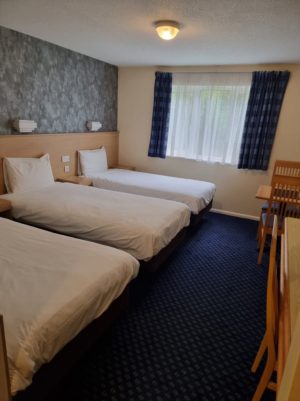 Habitación de hotel con tres camas, cabecera de madera, ventana con cortinas y una pequeña mesa.