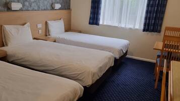 Habitación de hotel con tres camas, cabecera de madera, ventana con cortinas y una pequeña mesa.